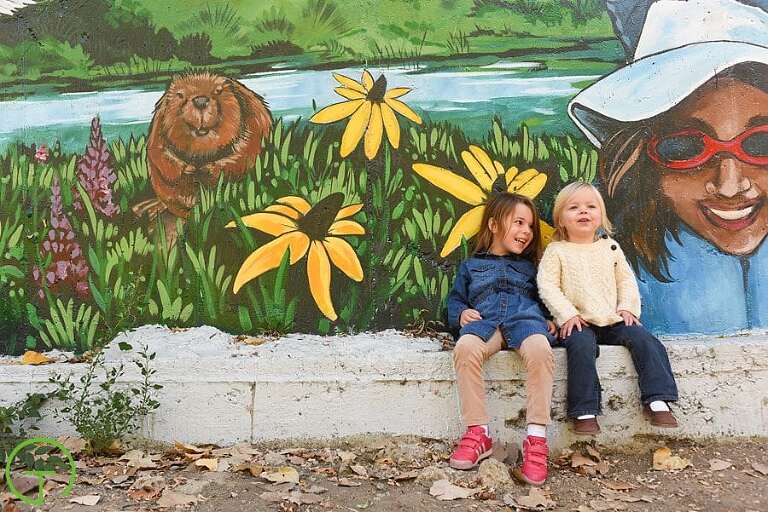 Two young sisters sit on a ledge with a colorful mural near the Huron River in Ypsilanti's Depot Town.