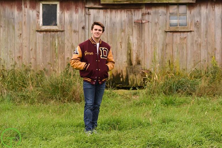 A high school senior poses with his varsity jacket near a rustic barn for his ypsilanti senior portraits