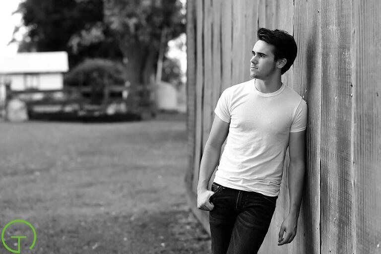 A high school senior leans against a rustic barn during his senior portrait session