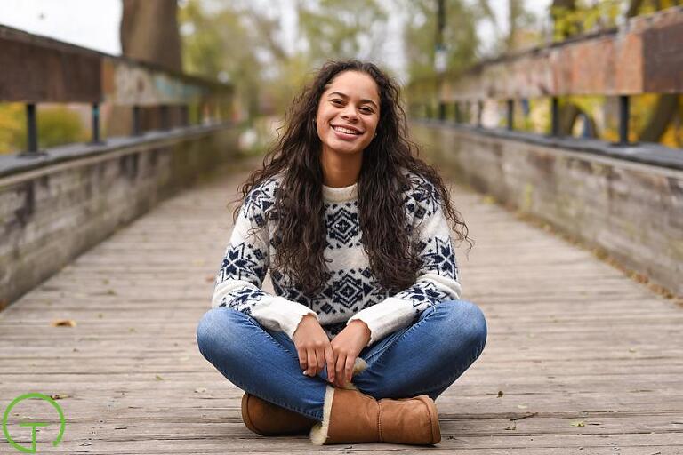 A high school senior casually sits on a wooden bridge in Ypsilanti’s Depot Town during her Ypsilanti senior portrait session