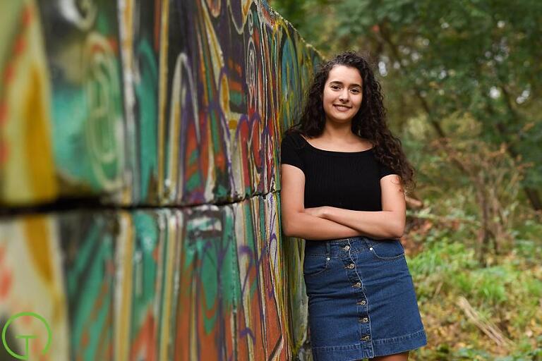 A teenager leans against a graffiti wall in ann arbor surrounded by lush green trees