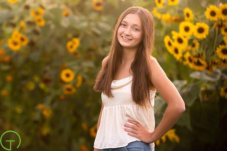 A high school senior girl stands near a sunflower field for her senior portrait