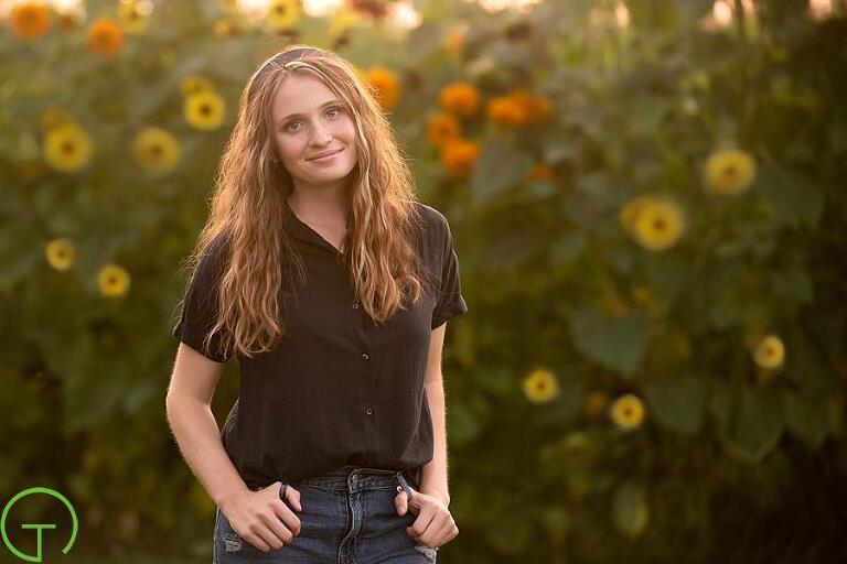 A high school senior girl stands in a sunflower field posing for a senior picture