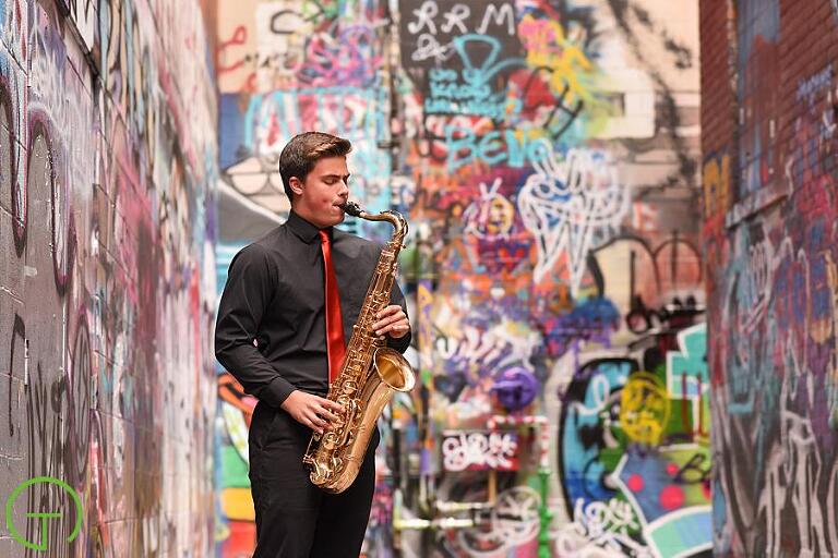 A teenage boy plays saxaphone in an alley painted with colorful grafitti 