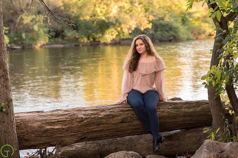 a high school girl sits on a log along the huron river with the sun setting behind her