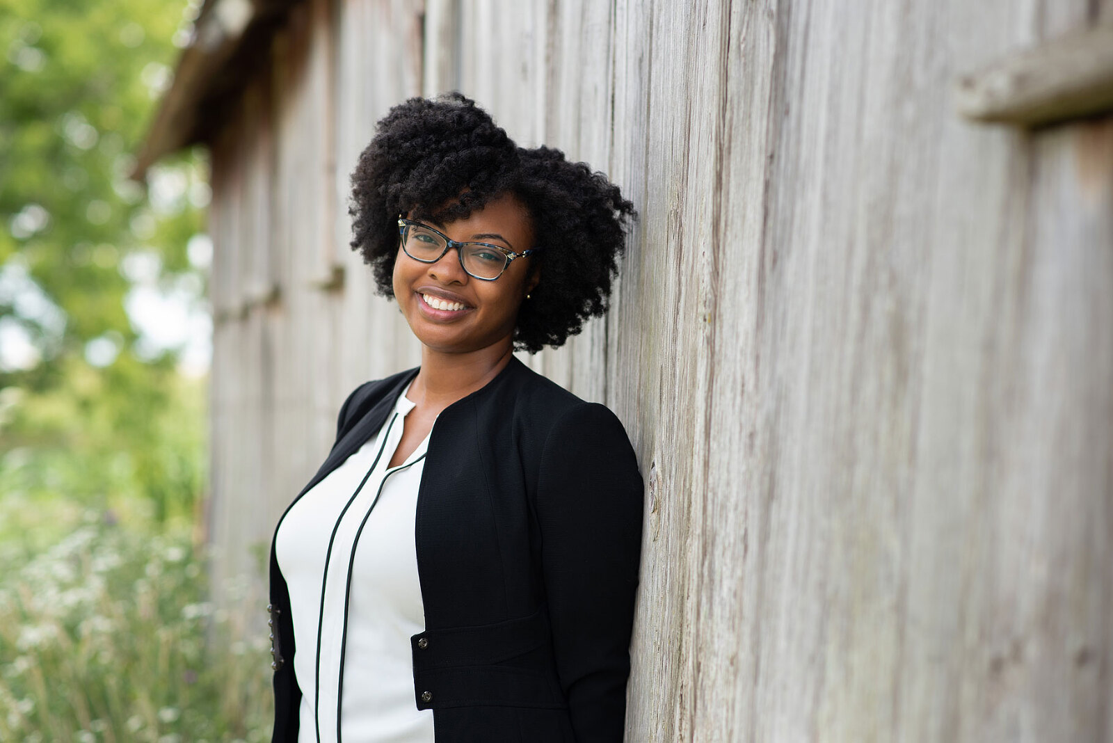 a young professional poses near an old barn for a headshot portrait