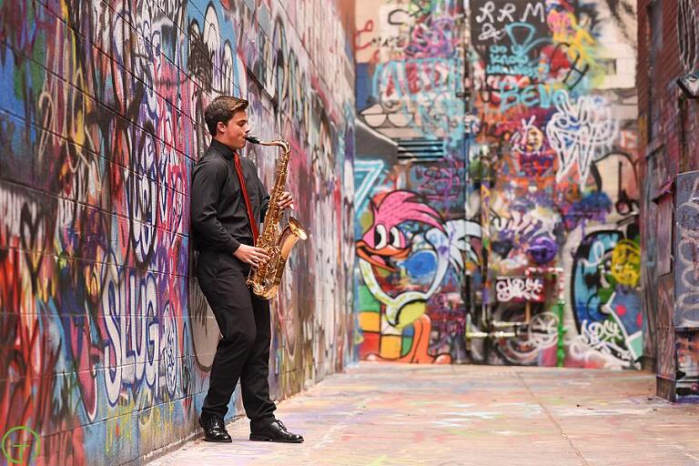 A high school senior boy stands in colorful graffiti alley playing his saxophone