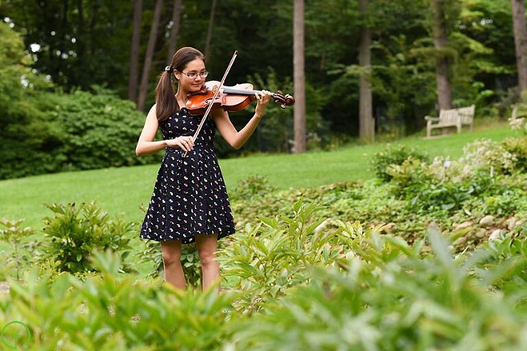 a high school senior girl plays her violin in a lush green setting in nichols arboretum